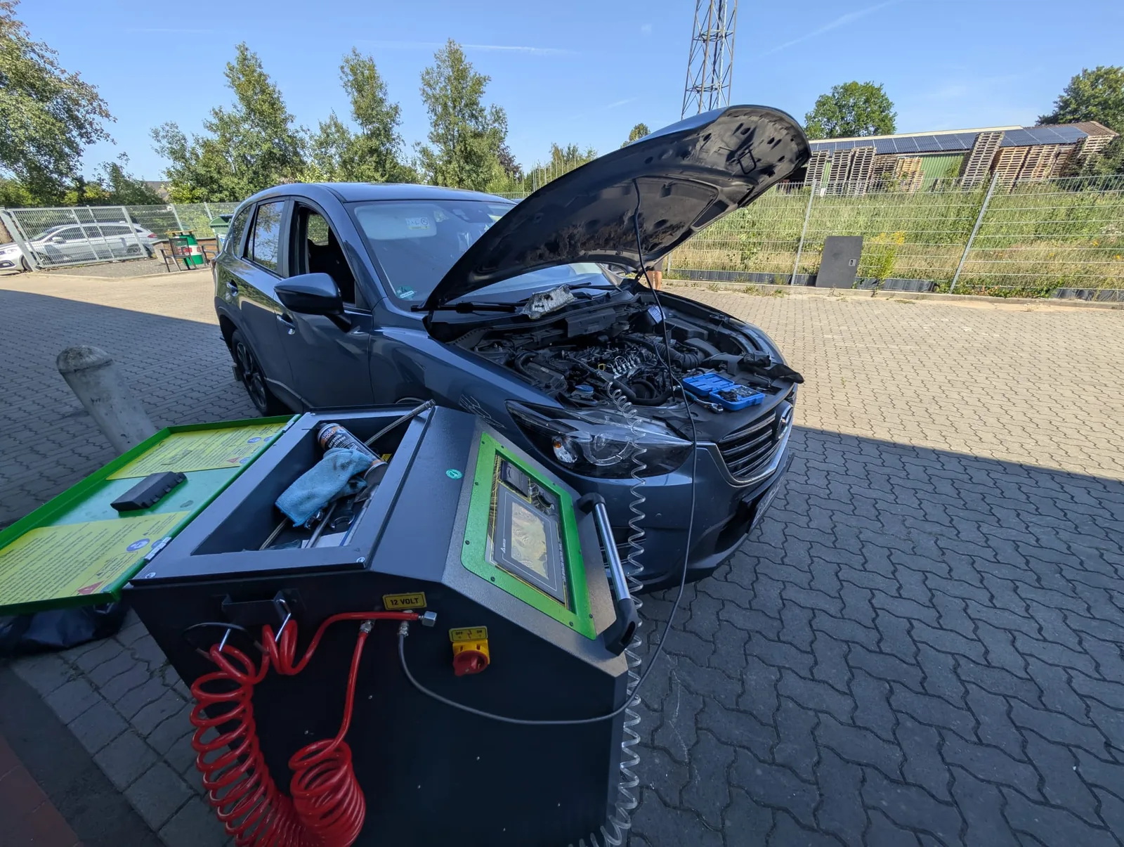 Grauer Mazda mit geöffneter Motorhaube auf dem Hof, angeschlossen an das grüne mobile Reinigungsgerät